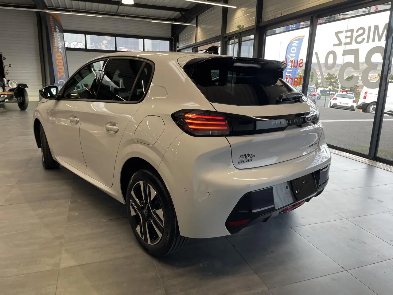 Vue 3/4 arrière droite d'une Peugeot 208 blanche Okenite avec bandeau noir brillant et feux arrière à LED en intérieur showroom.