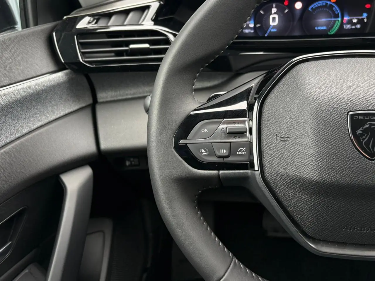 Gros plan sur le volant noir de la Peugeot 308 SW Hybrid 2025 avec commandes intégrées et tableau de bord numérique partiel.