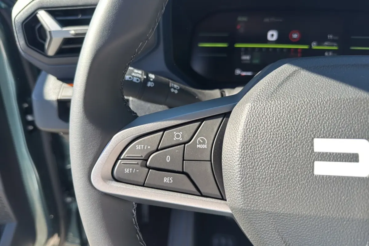Gros plan sur les commandes au volant côté gauche du Dacia Bigster 2025 vert cèdre, avec affichage numérique en arrière-plan.