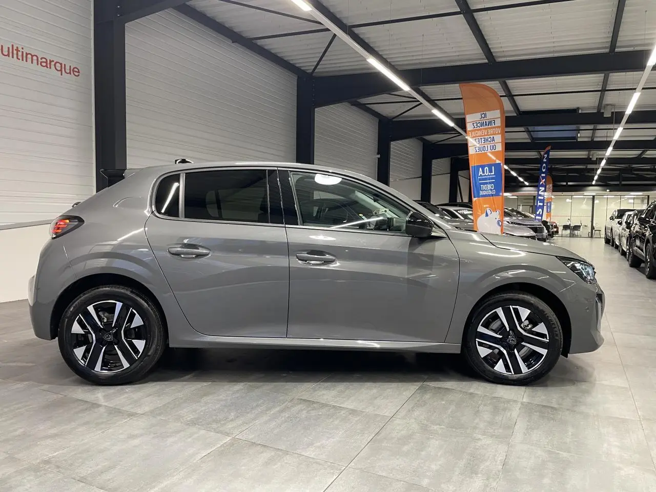 Profil côté gauche d'une Peugeot 208 gris sélénium 2025 avec jantes alliage bi-ton dans un showroom.