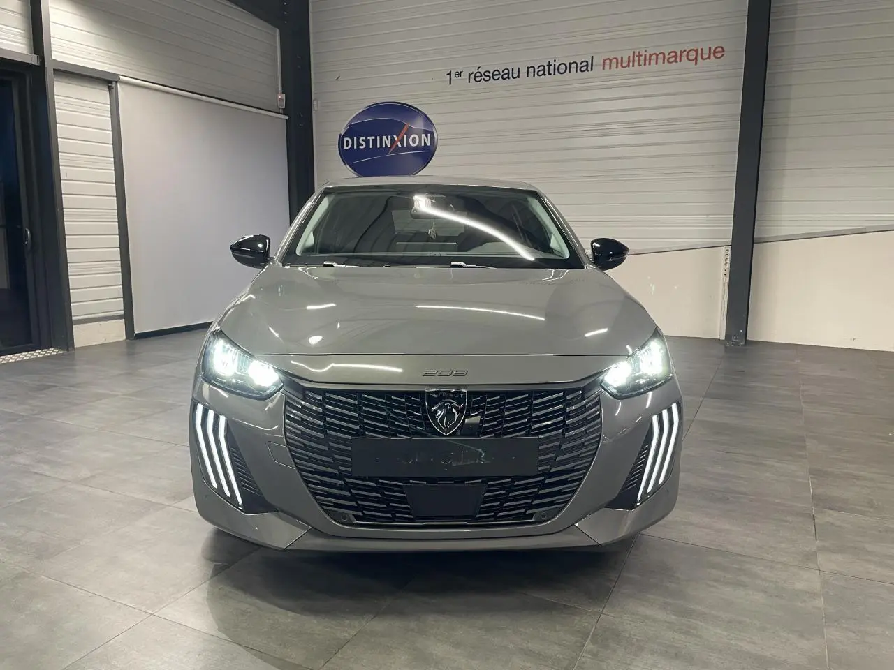 Vue avant d'une Peugeot 208 gris selenium avec phares LED allumés et calandre noire brillante en intérieur showroom.