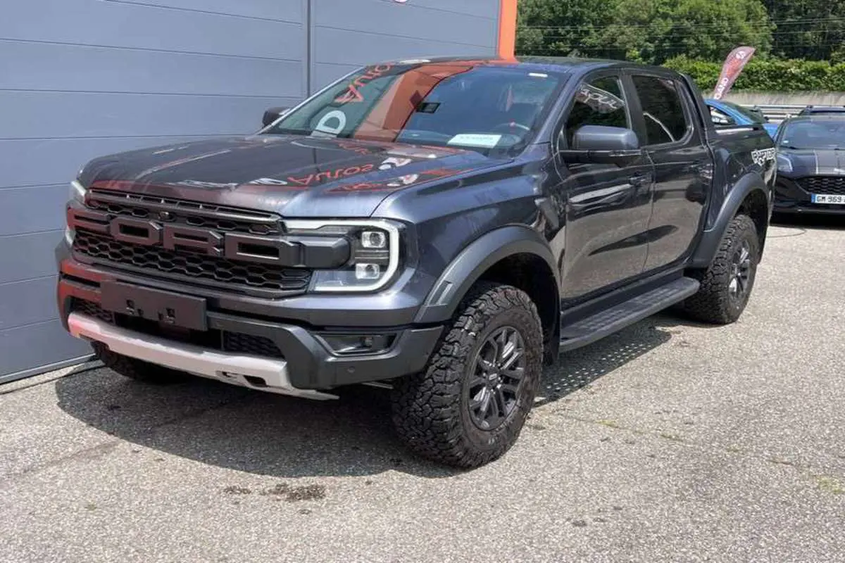 Ford Ranger Raptor gris météore en 3/4 avant droit, avec calandre imposante et pneus tout-terrain marqués.