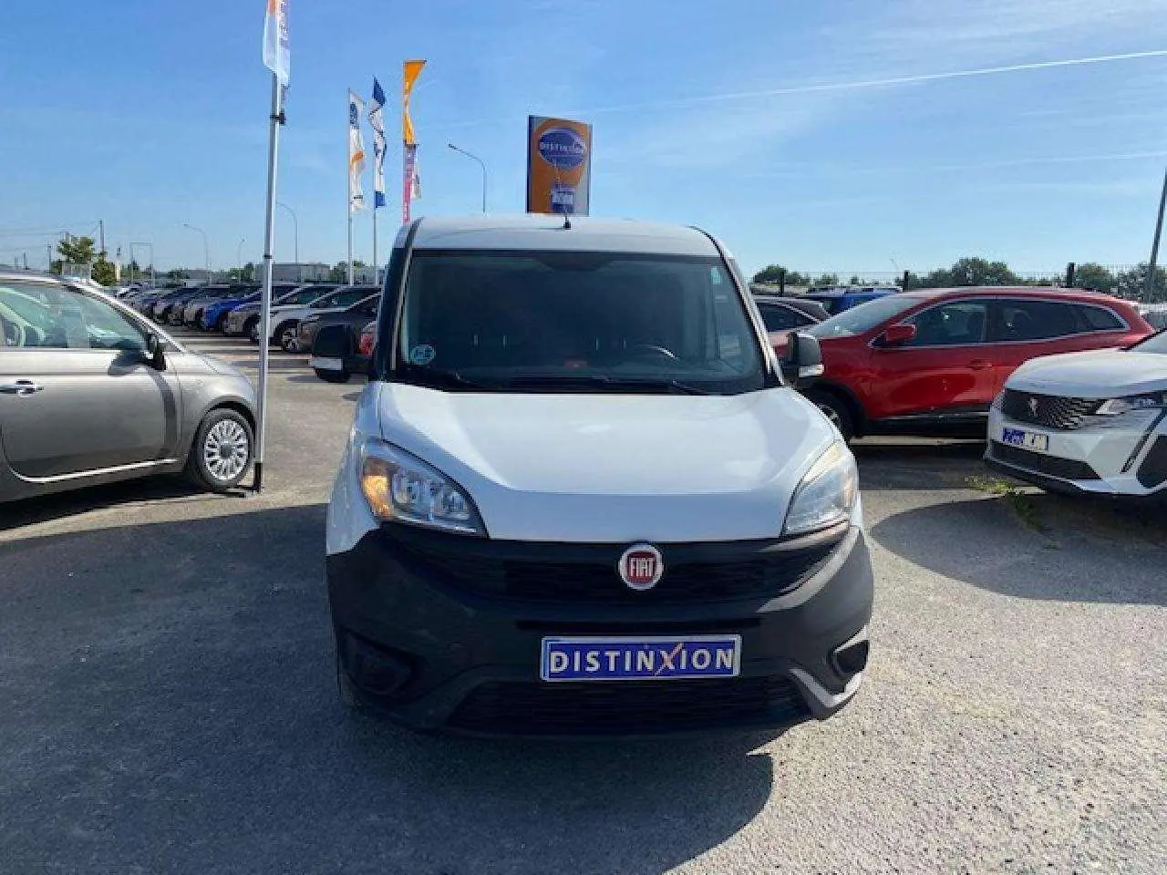Vue avant d'un FIAT Doblo blanc avec pare-chocs noir mat, stationné sur un parking en plein jour.