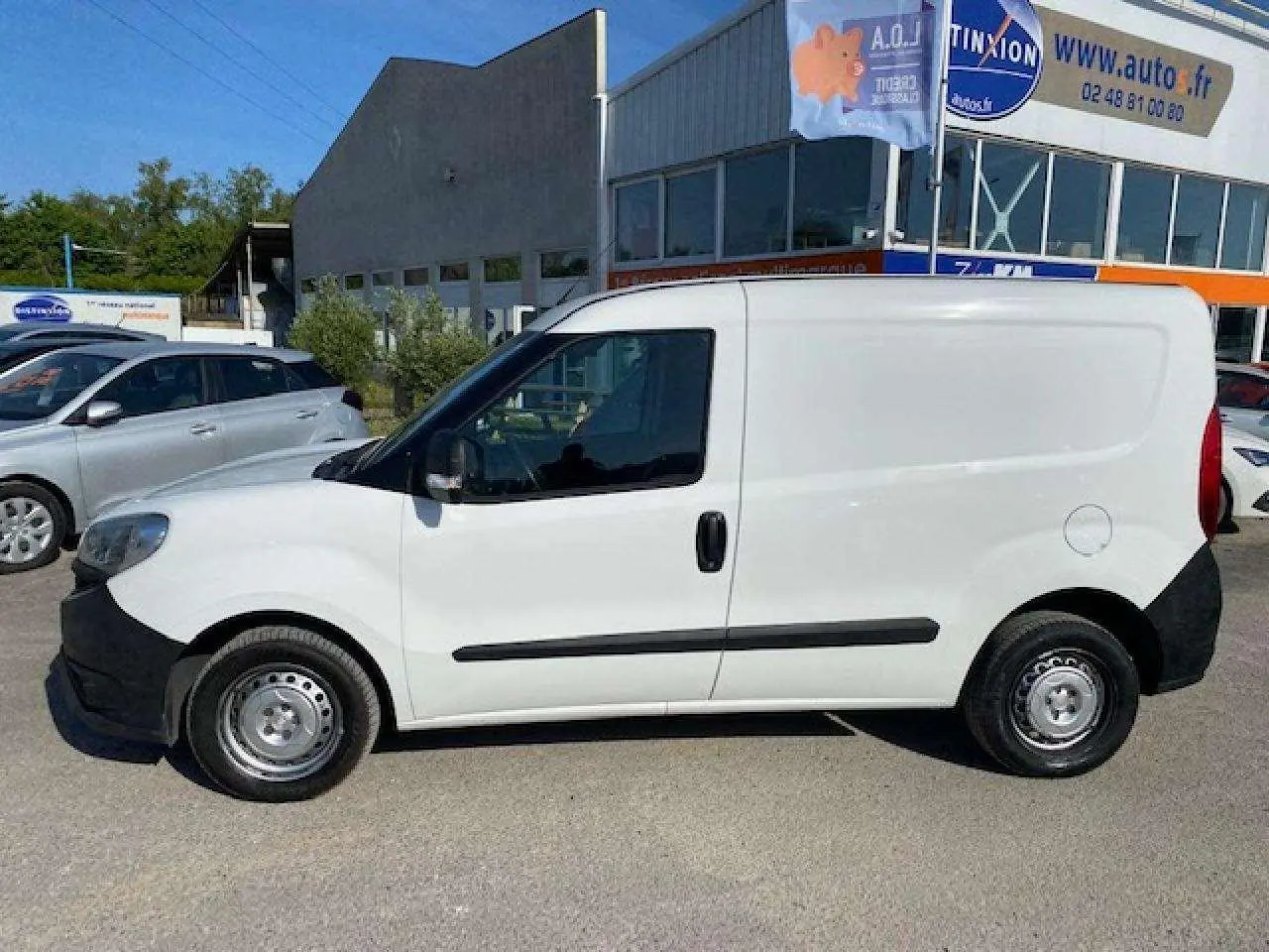 Vue de profil côté gauche d’un FIAT Doblo blanc fourgon tôlé avec pare-chocs noirs et porte latérale droite visible en arrière.