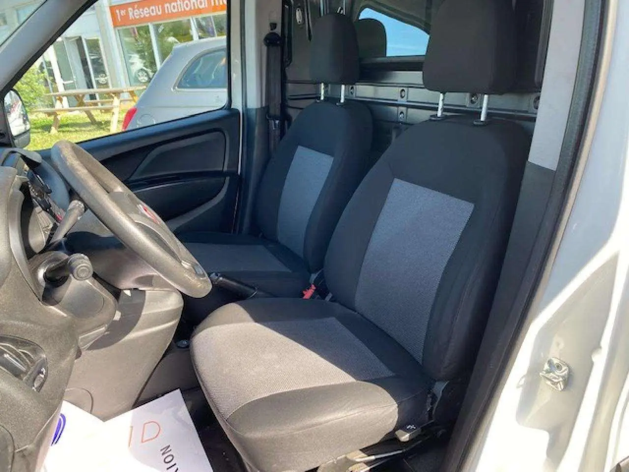 Vue intérieure côté conducteur du Fiat Doblo blanc 2017, montrant les sièges avant et la paroi de séparation vitrée.