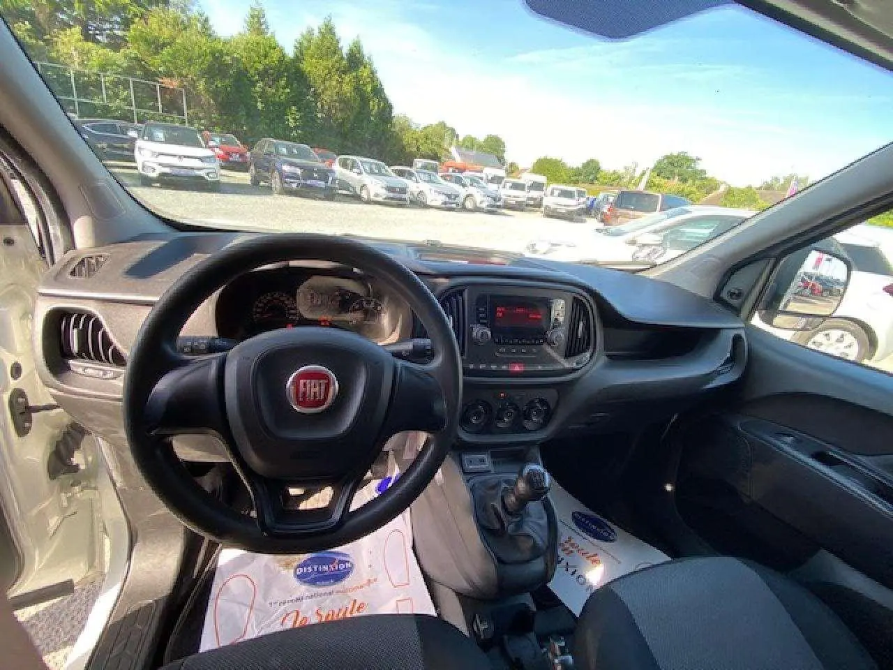 Vue intérieure du poste de conduite du Fiat Doblo blanc de 2017, avec volant, tableau de bord et levier de vitesse manuelle visibles.