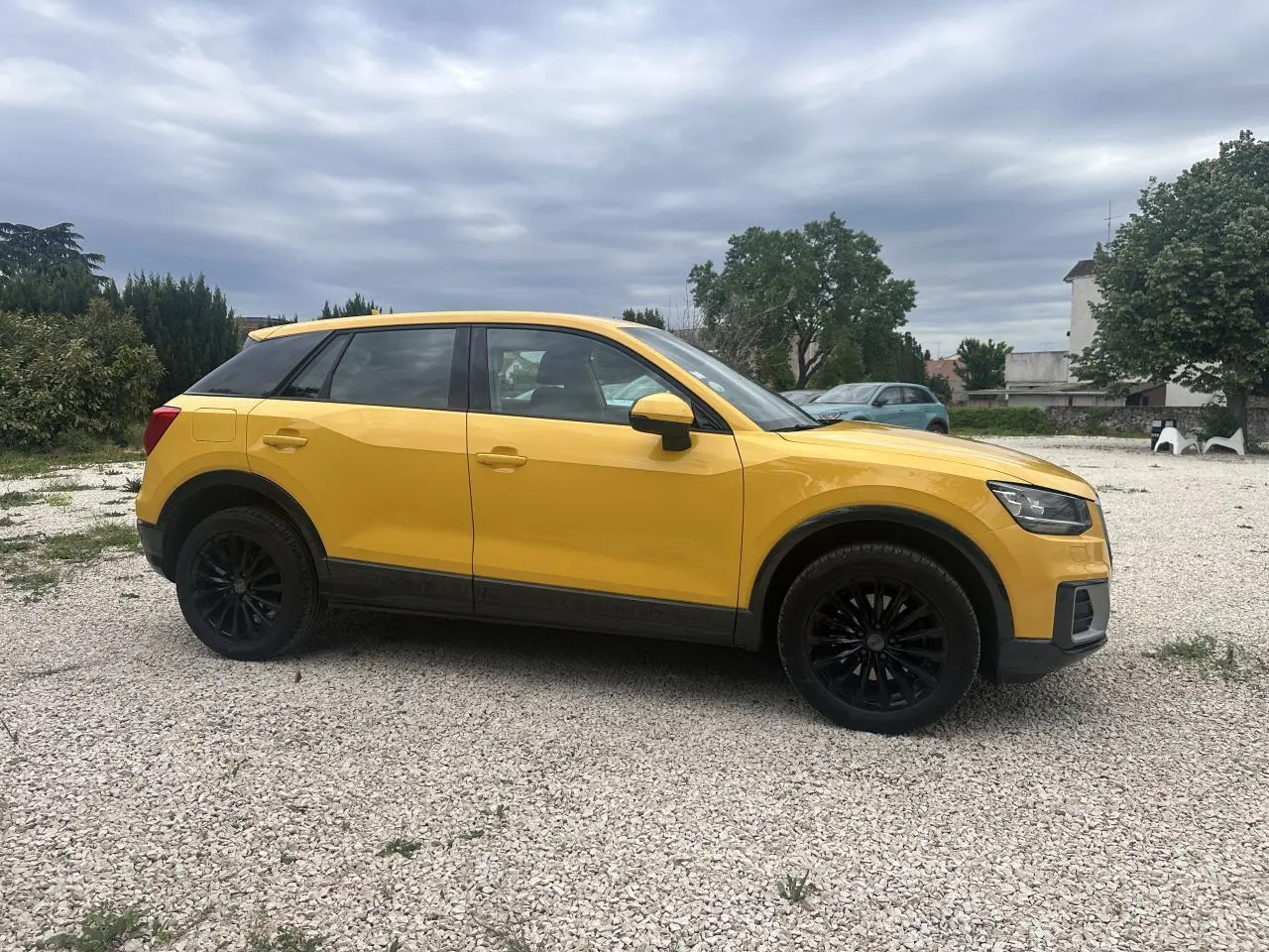 Audi Q2 jaune vue de profil côté gauche avec jantes noires et toit noir sur sol gravillonné.