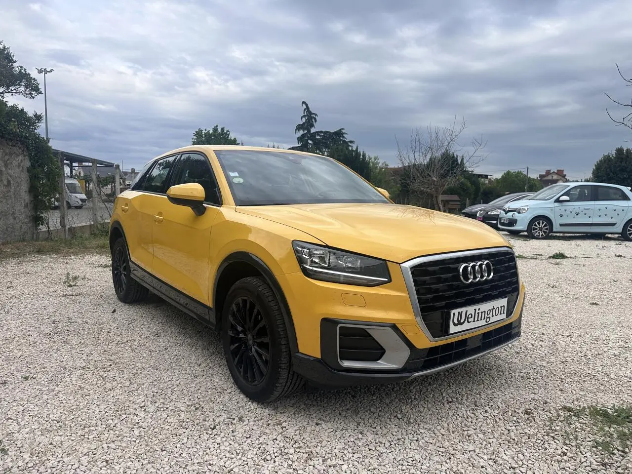 Audi Q2 jaune vue 3/4 avant droit avec calandre noire et jantes noires sur parking gravillonné.