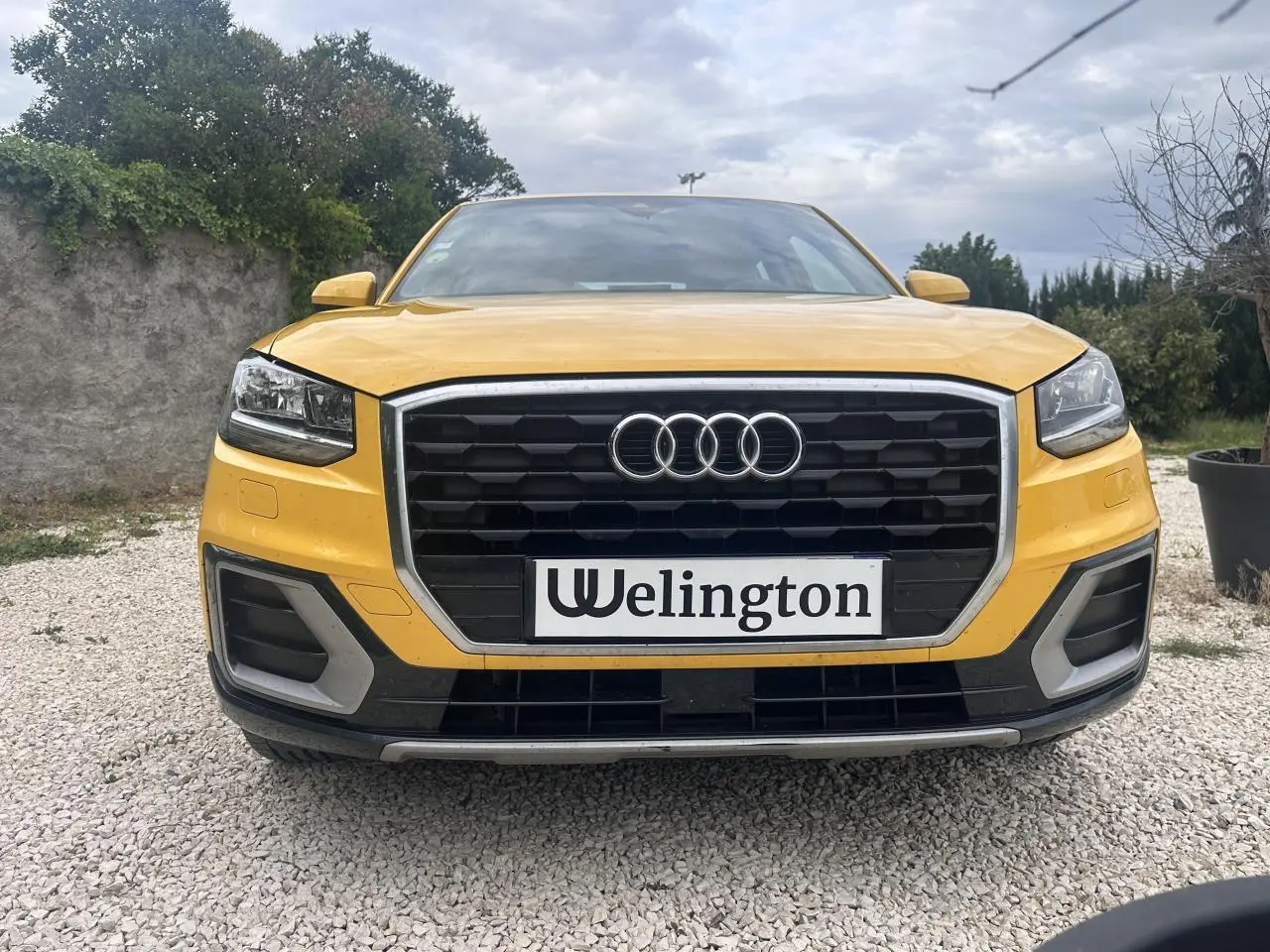 Vue frontale d'un Audi Q2 jaune avec calandre noire et contours chromés, sur sol gravillonné.