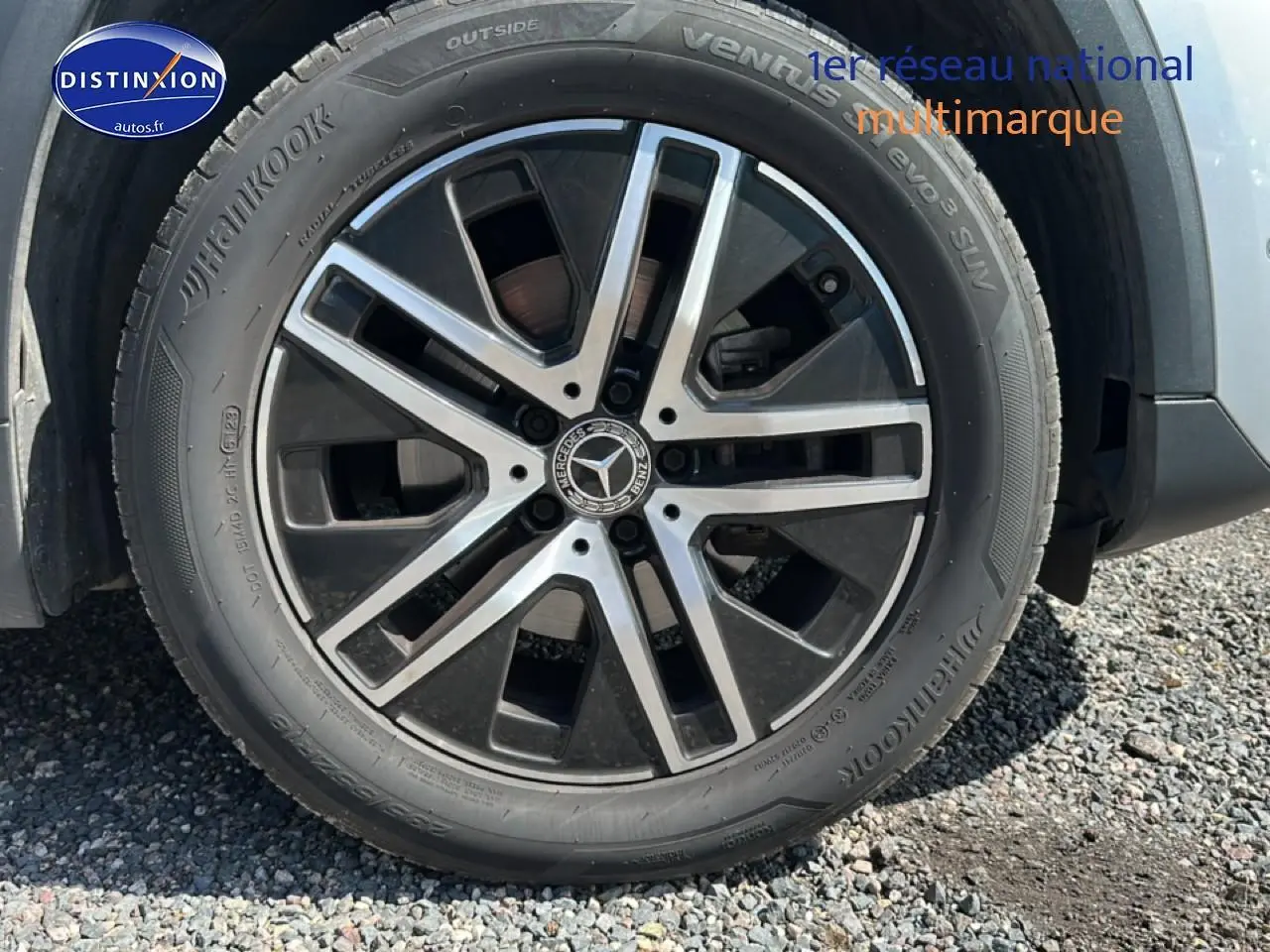 Gros plan sur la jante alu noire et argentée d’un Mercedes GLA gris, avec pneu Hankook visible sur gravier.