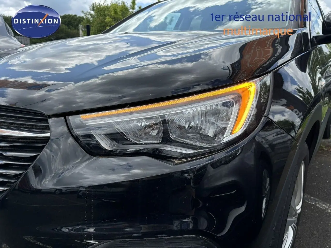 Gros plan sur le phare avant droit d’un Opel Grandland X noir brillant, reflétant le ciel et les environs.