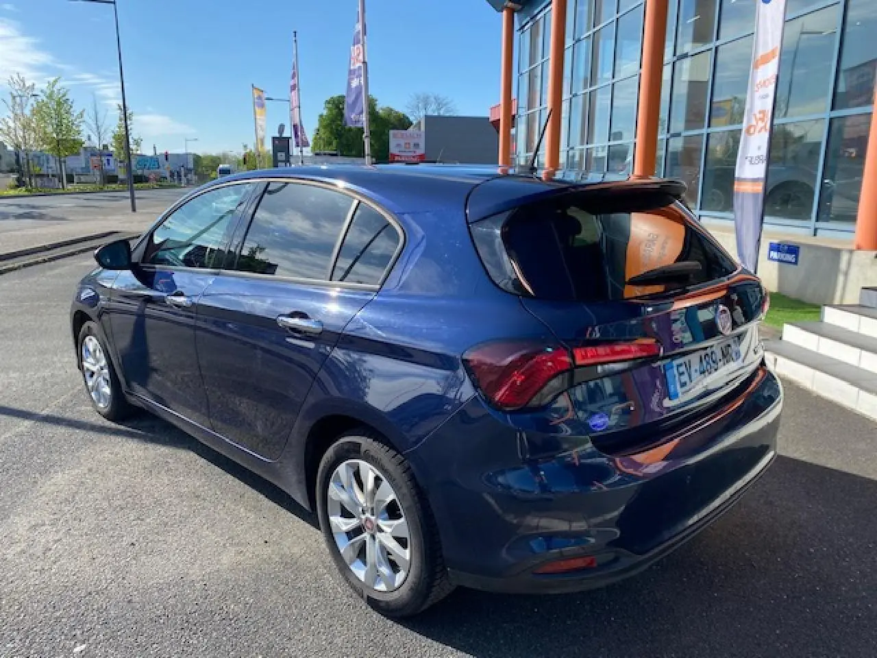 Vue 3/4 arrière droite d'une Fiat Tipo bleu métallisé garée devant un bâtiment avec vitres surteintées et jantes argentées.