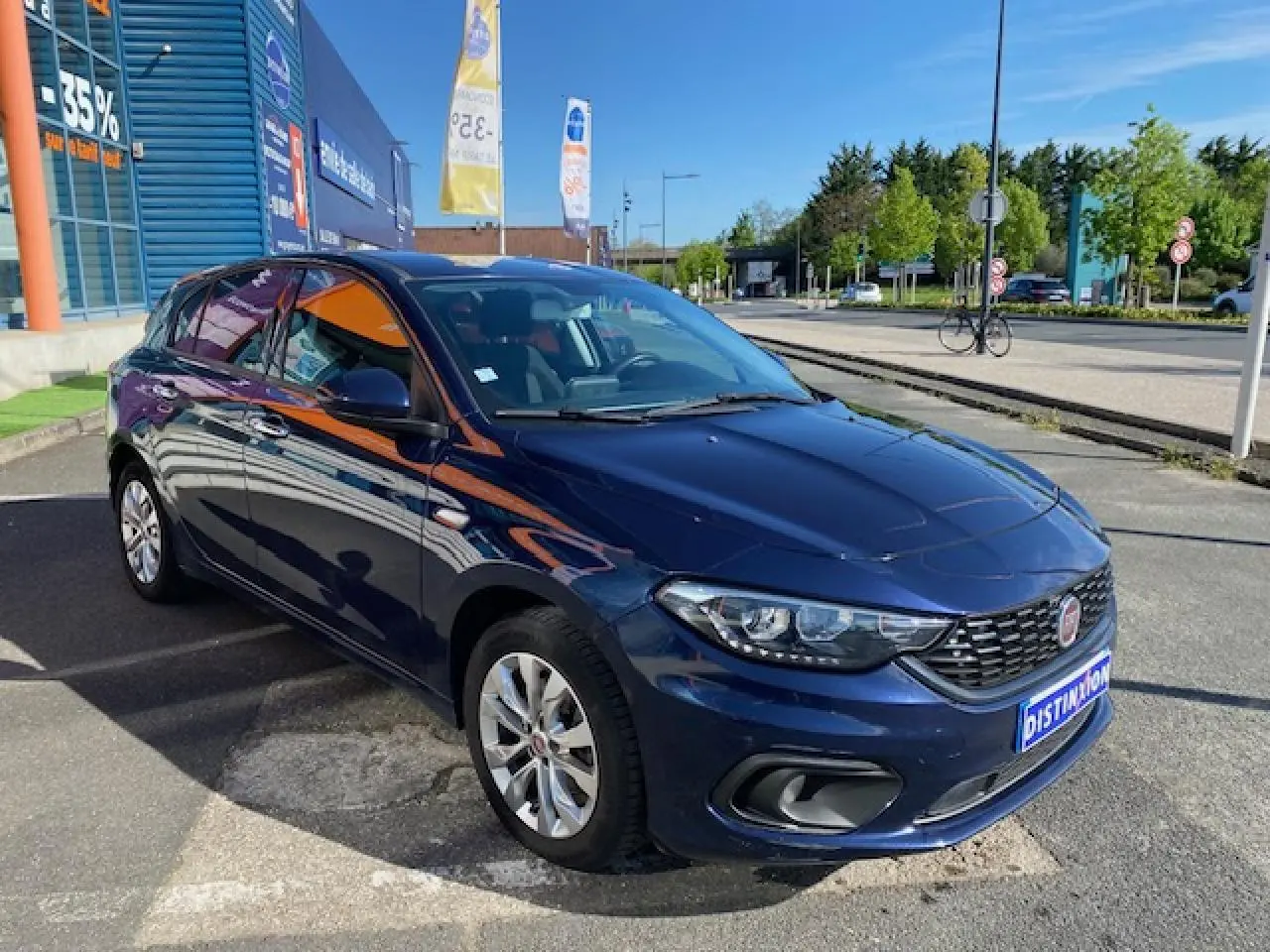 Vue 3/4 avant droite d'une FIAT Tipo bleu métallisé garée devant un bâtiment commercial par temps ensoleillé.