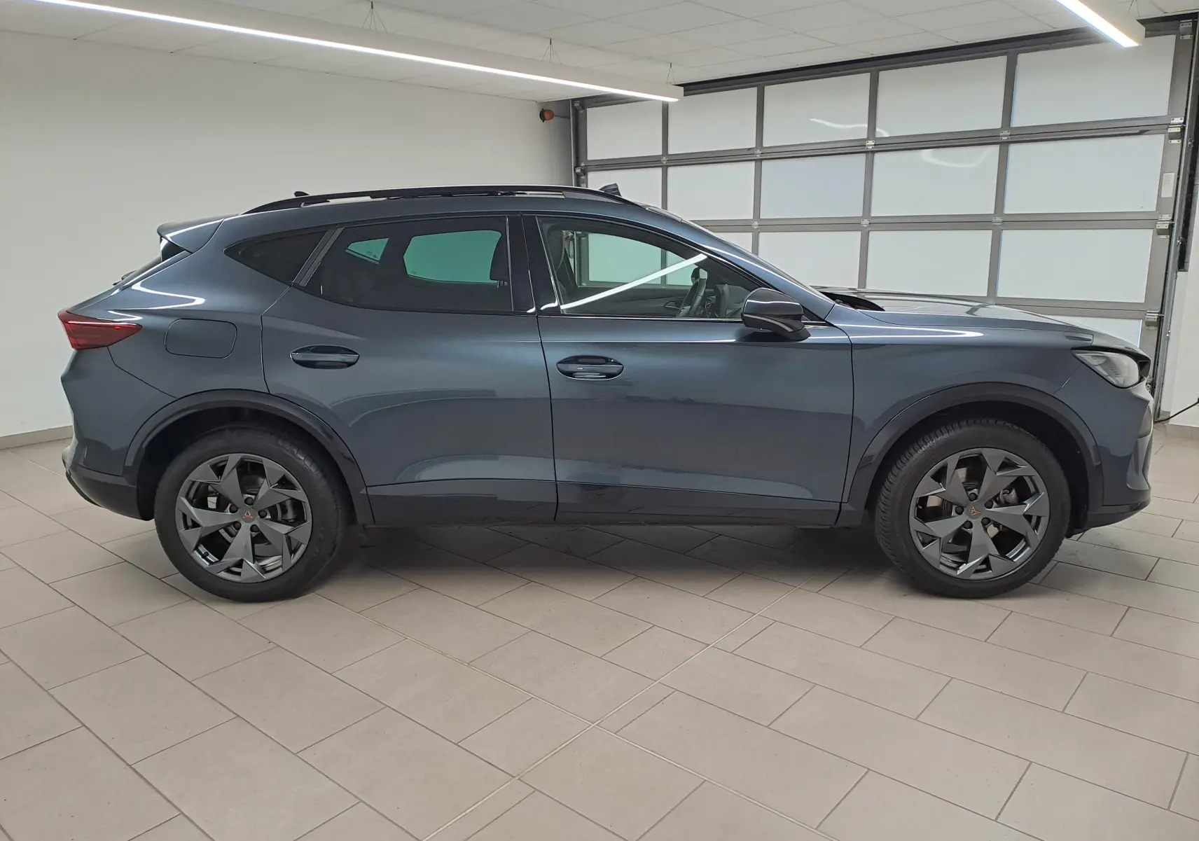 Vue de profil côté gauche d'un CUPRA Formentor gris magnétique avec jantes alliage et toit ouvrant panoramique en intérieur showroom.