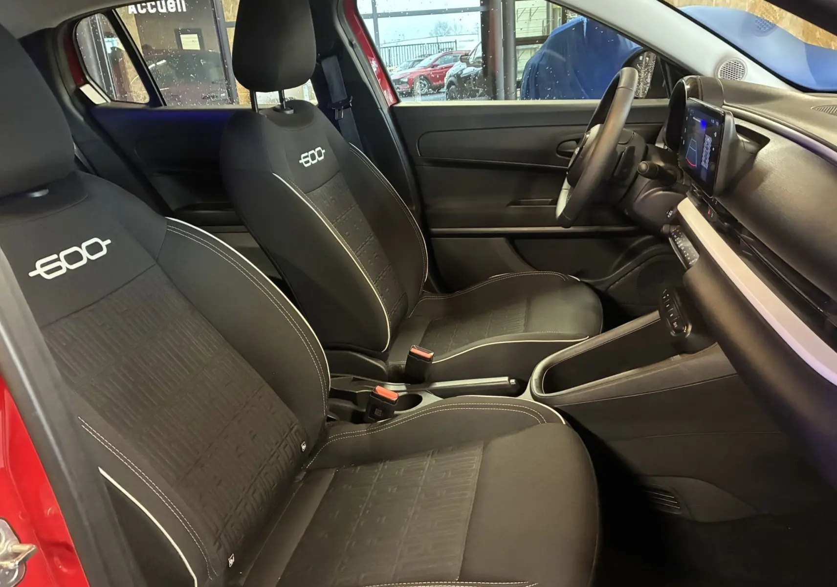Vue intérieure côté conducteur de la FIAT 600 rouge, sièges noirs avec logo 600 brodé et tableau de bord moderne.