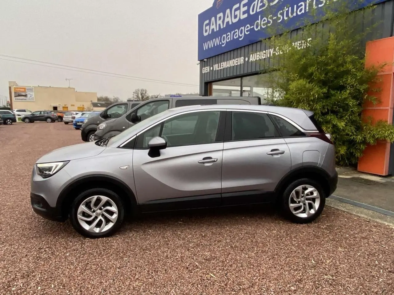 Opel Crossland X gris métallisé en profil droit, avec jantes alliage 16'' et vitres surteintées, stationné devant un garage.