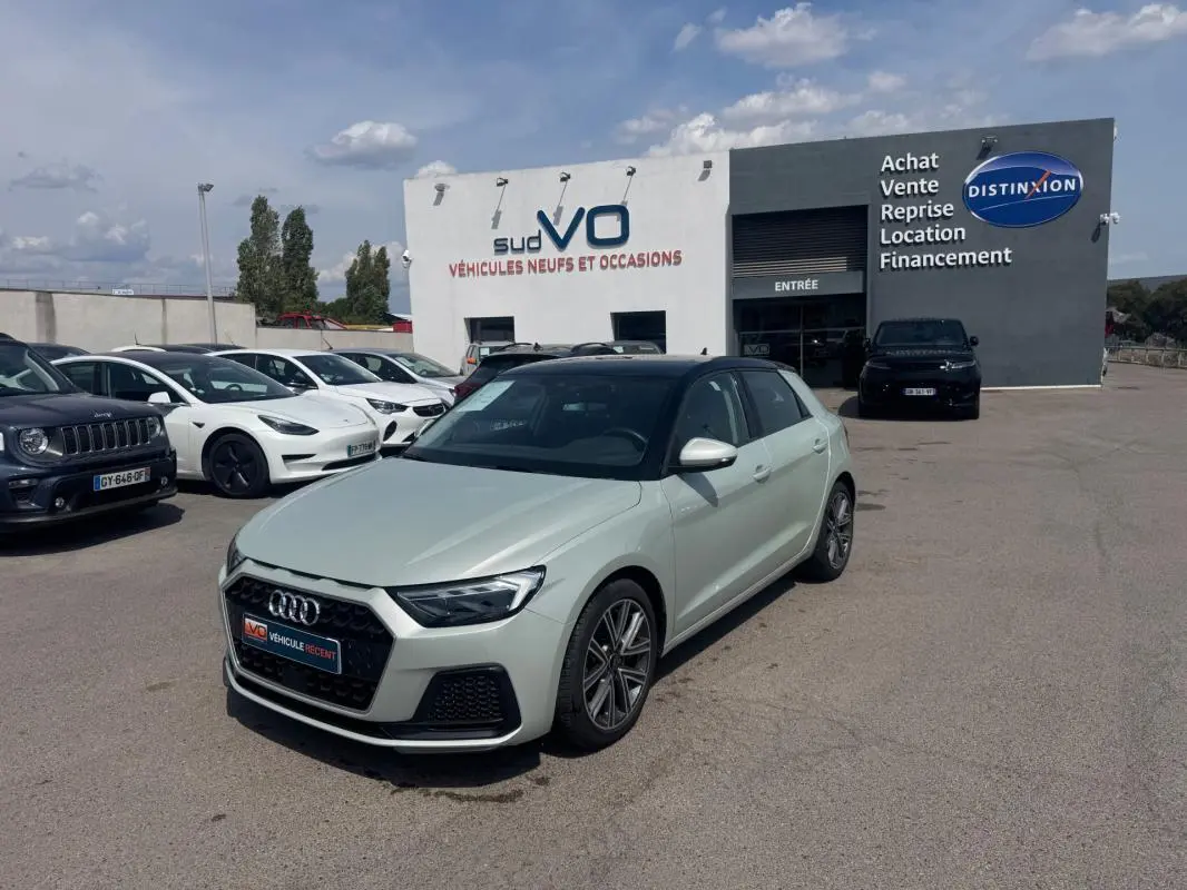 Audi A1 Sportback gris vue 3/4 avant droit, avec toit noir, stationnée devant un garage de véhicules neufs et occasions.