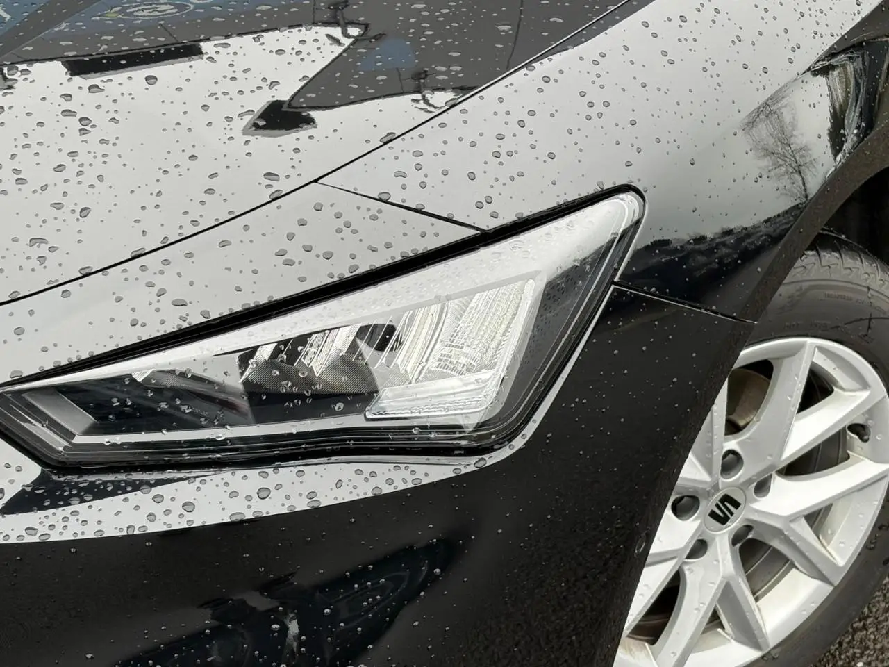 Gros plan sur le phare avant droit et la jante alliage 16 pouces d'une SEAT LEON 2.0 TDI noir Minuit sous la pluie.