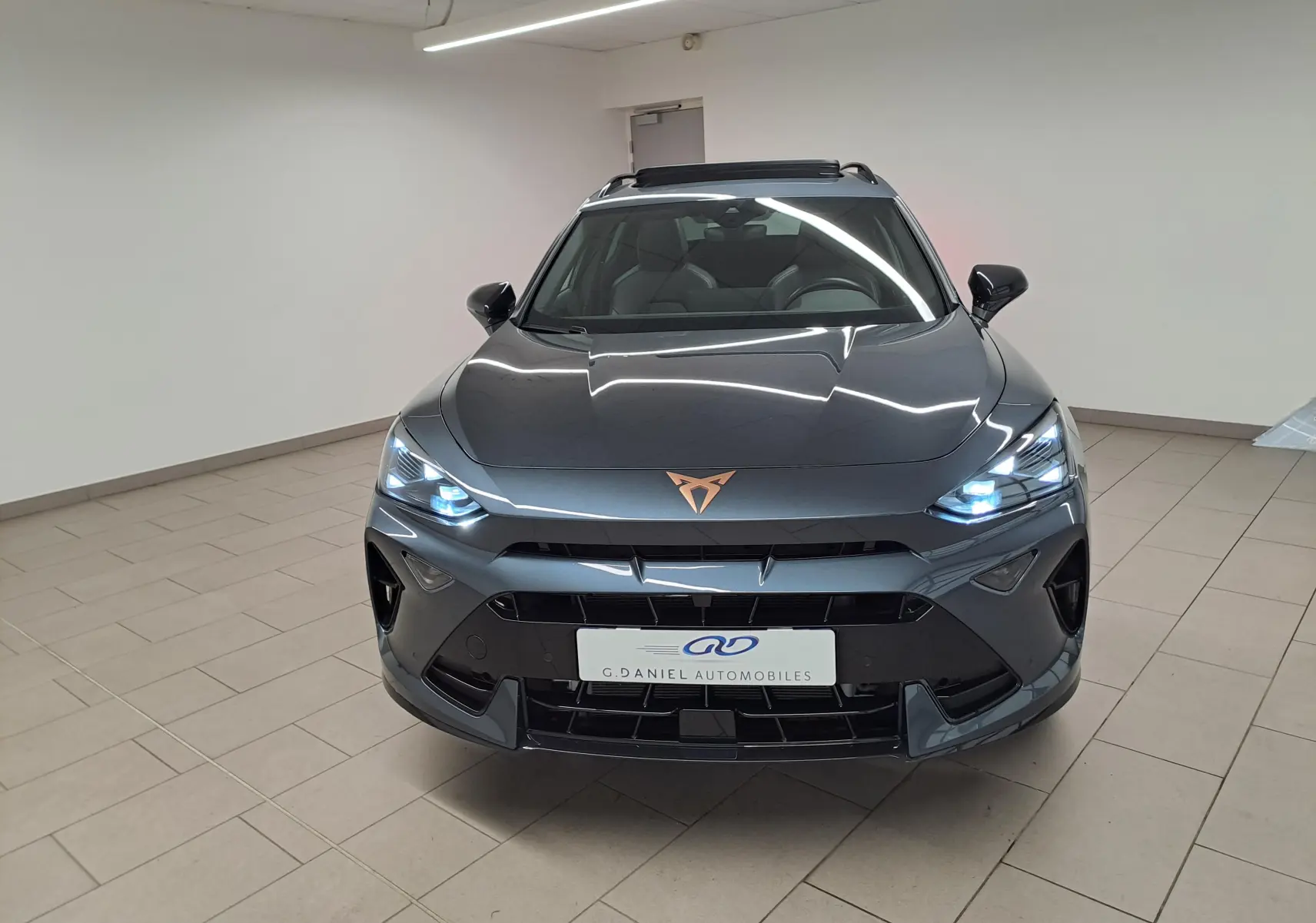Vue frontale d'un CUPRA Formentor gris magnétique avec logo cuivré et phares LED allumés dans un showroom.