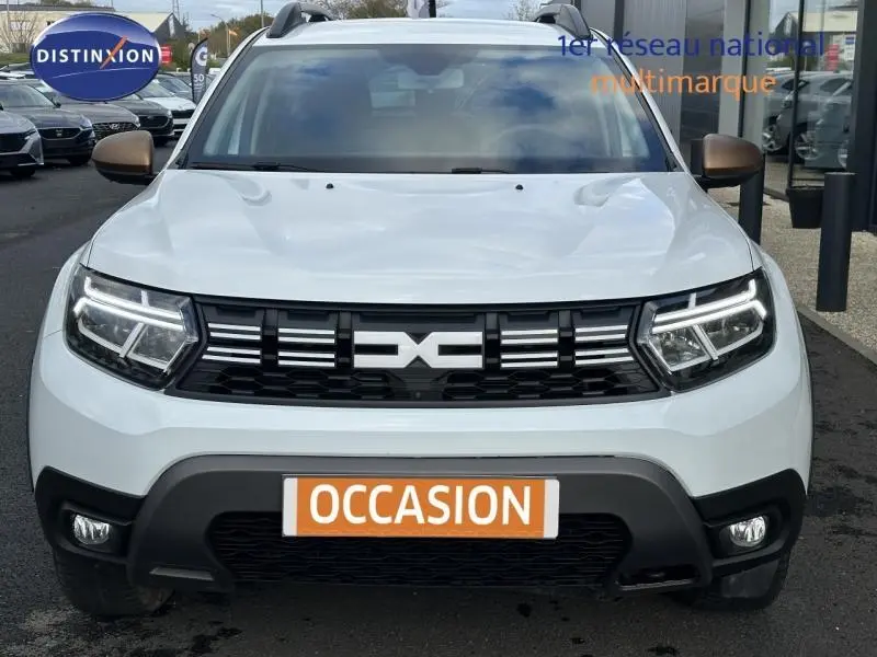 Vue frontale d'un Dacia Duster blanc Glacier 2024 avec phares LED allumés et calandre noire marquée du logo DACIA.