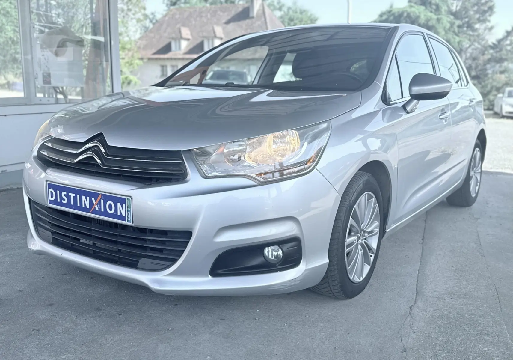 Citroën C4 gris vue 3/4 avant droit avec phares allumés et jantes alliage, stationnée devant un bâtiment.