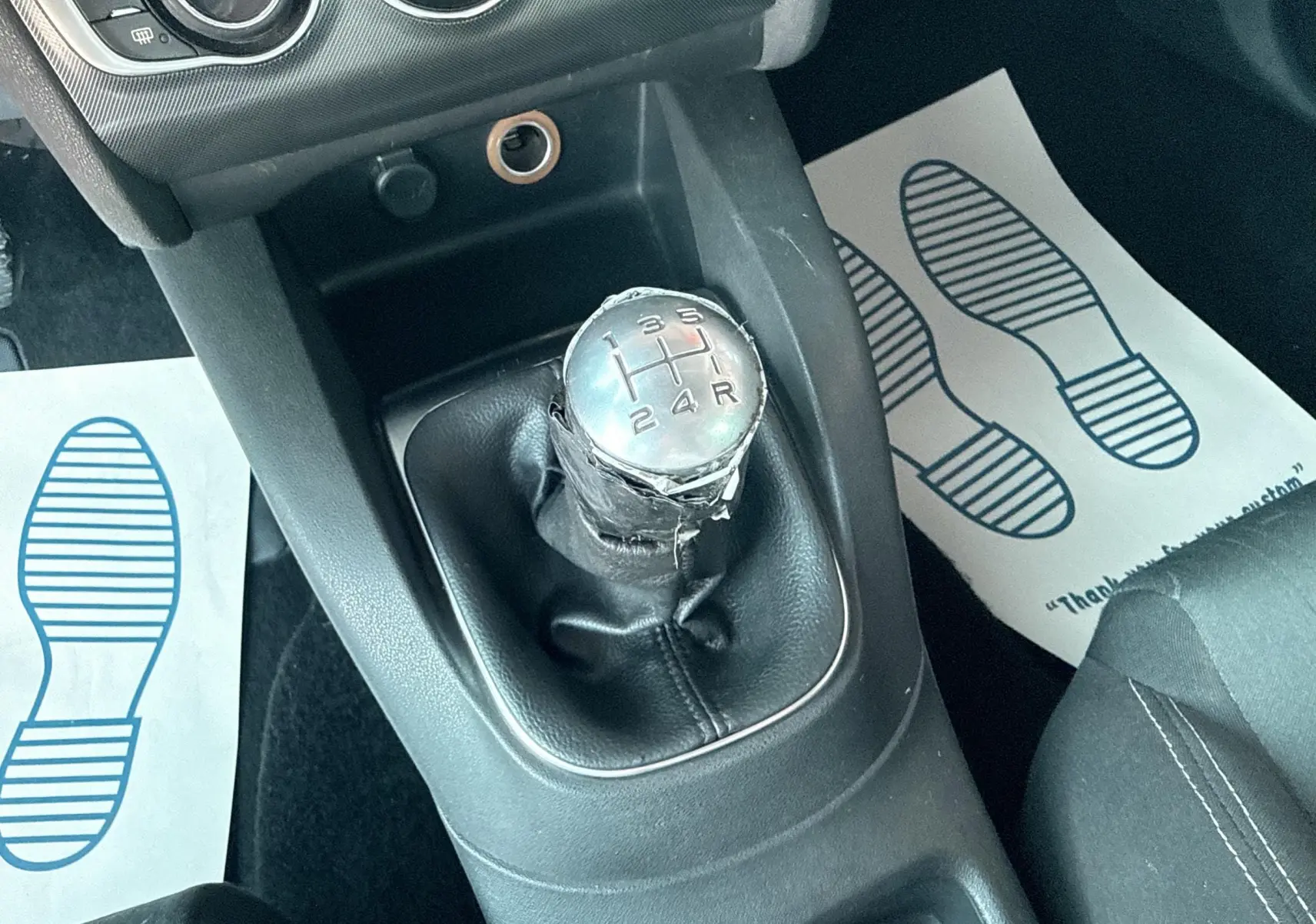 Gros plan sur le levier de vitesse manuel en cuir noir de la Citroën C4 gris, avec console centrale et tapis de sol visibles.