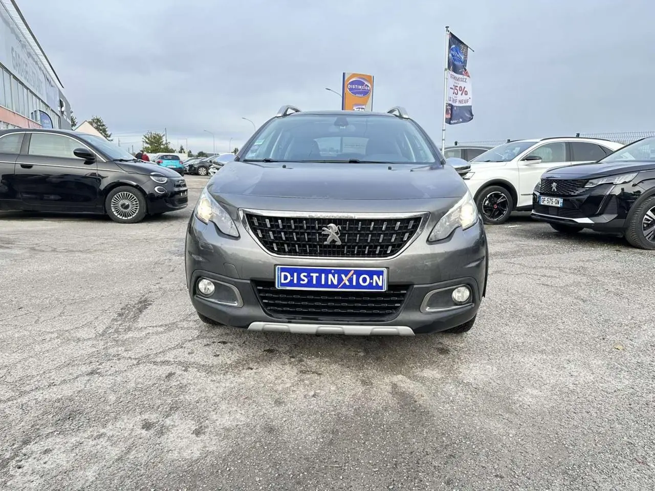Vue frontale d'un Peugeot 2008 gris foncé Crossway avec calandre noire et feux allumés sur parking extérieur.