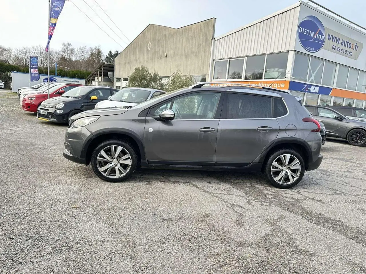 Profil latéral droit d'un Peugeot 2008 gris foncé avec jantes aluminium 17 pouces, stationné devant un bâtiment commercial.
