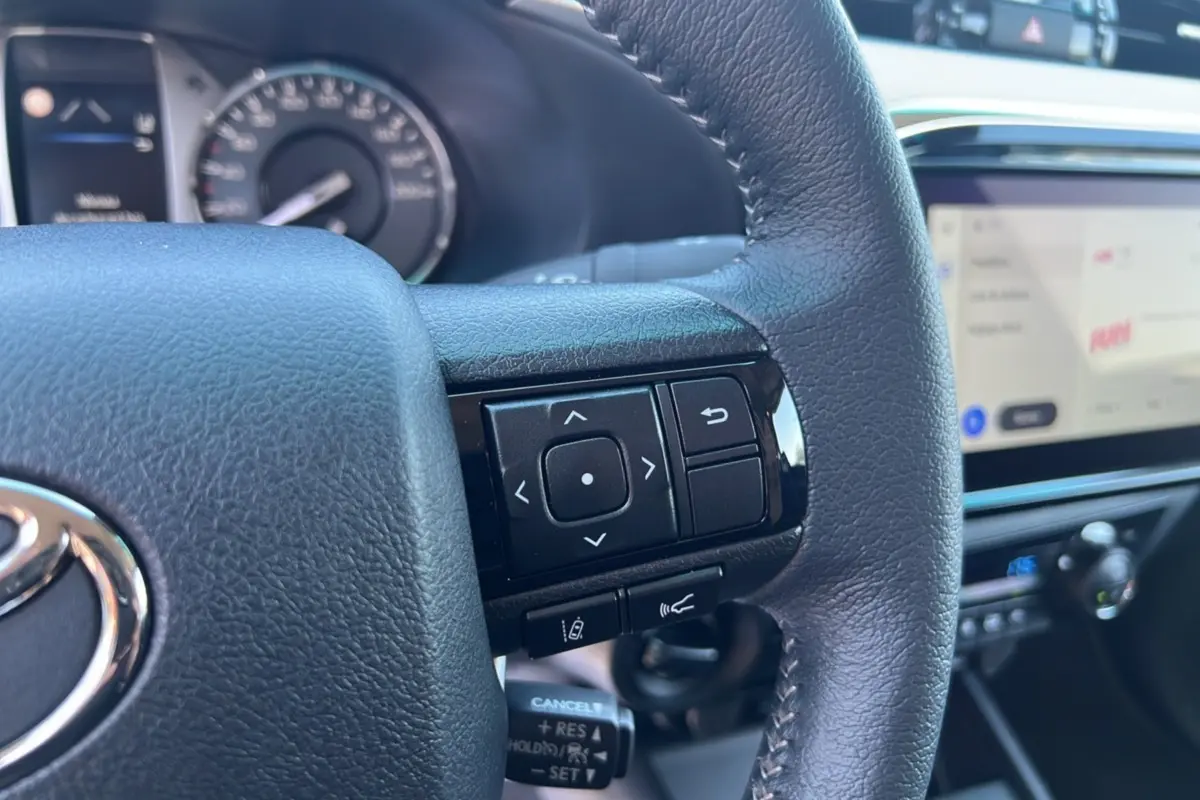 Gros plan sur le volant cuir multifonctions noir du Toyota Hilux 2026 avec écran tactile et tableau de bord flou en arrière-plan.