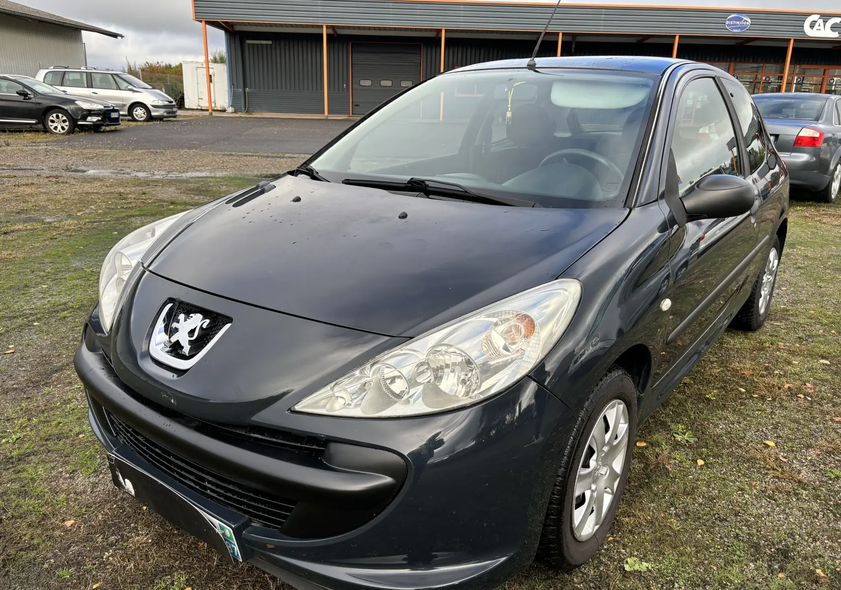 Peugeot 206+ gris foncé vue en 3/4 avant droit, avec phares larges et logo lion distinctif sur le capot.