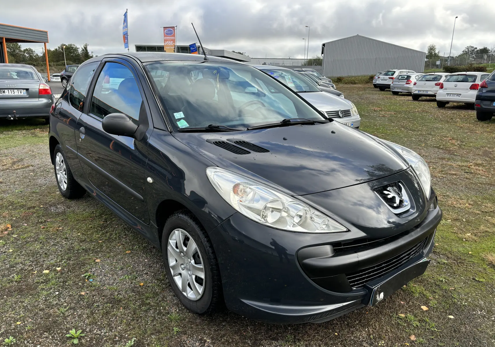 Peugeot 206+ gris foncé vue en 3/4 avant droit sur un parking extérieur avec plusieurs voitures en arrière-plan.