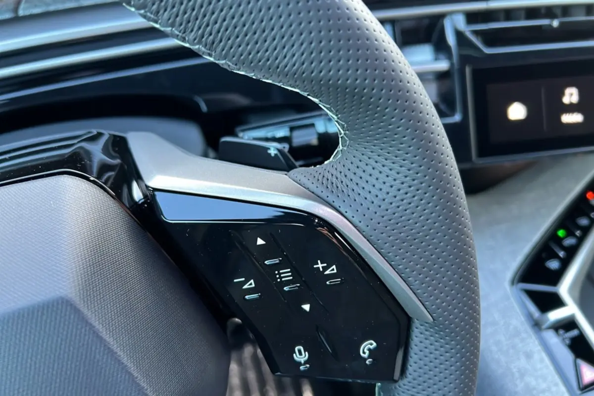 Gros plan sur le volant en cuir perforé gris avec commandes multifonctions, tableau de bord et écran tactile partiellement visibles.