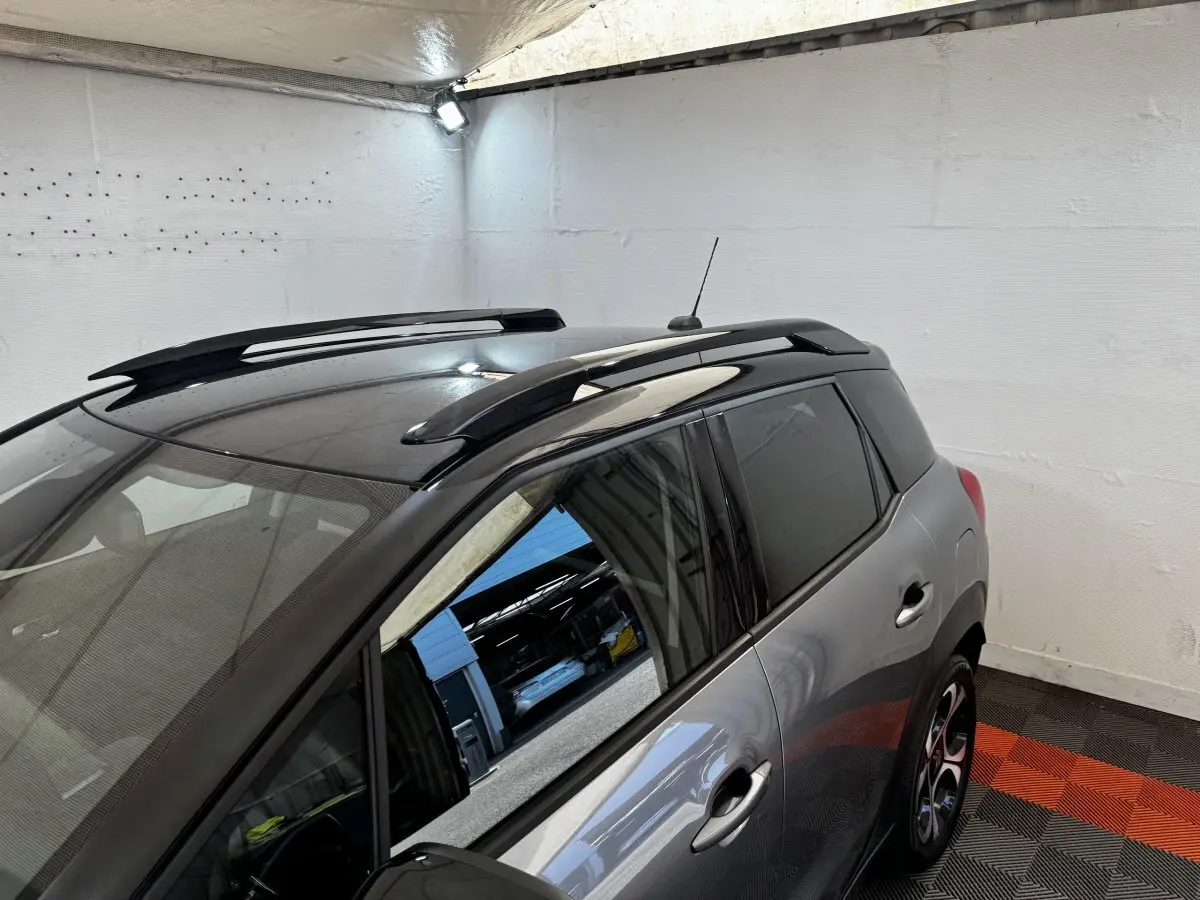 Vue 3/4 arrière droite d'une Citroën C3 gris foncé avec toit noir et barres de toit en intérieur fermé.