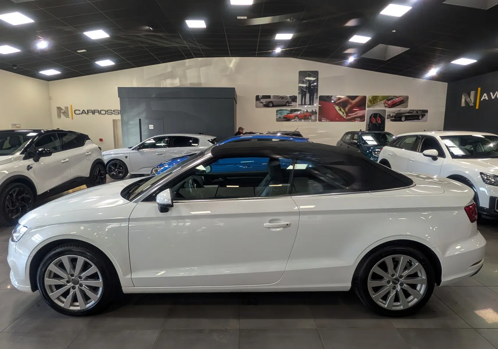 Audi A3 Cabriolet blanc vue de profil côté gauche, capote noire fermée, dans un showroom lumineux.