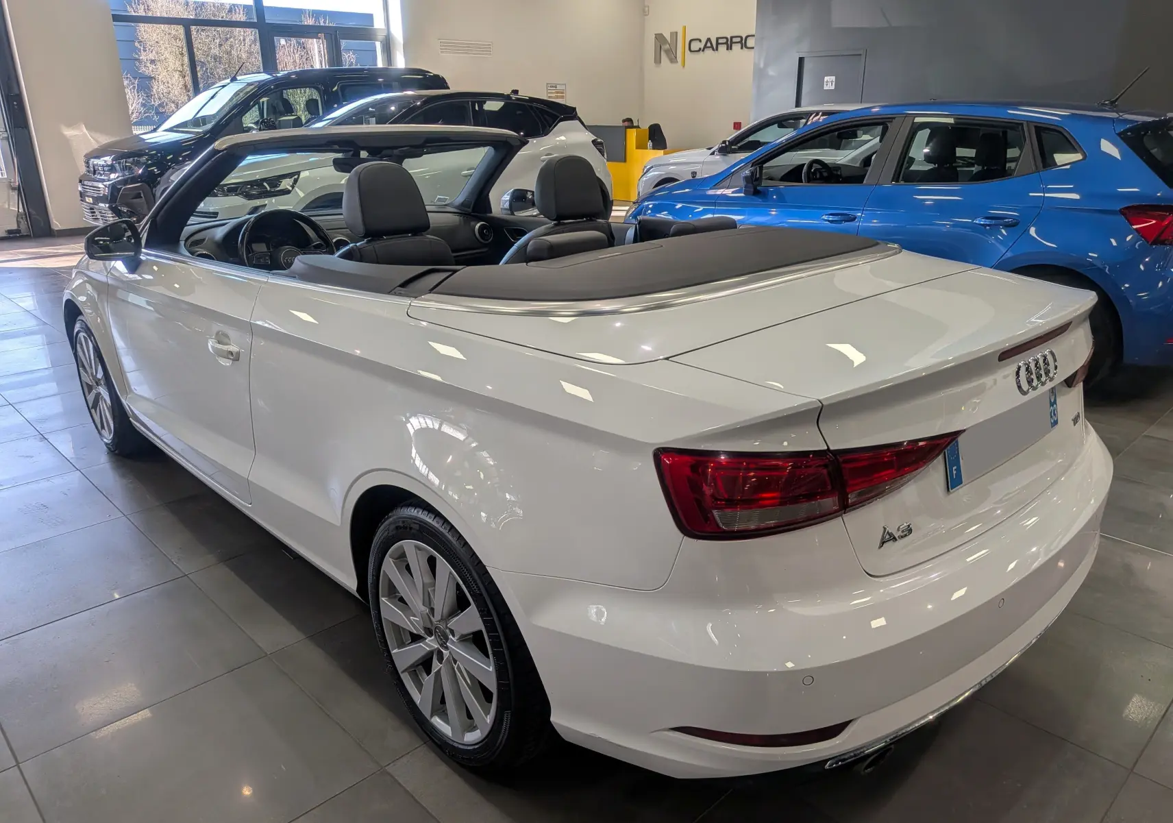 Audi A3 Cabriolet blanc vue 3/4 arrière droit, capote noire repliée, intérieur cuir noir visible en showroom.