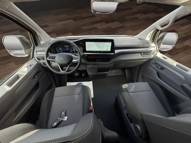 Vue intérieure avant du Volkswagen Transporter ProCab 2026, tableau de bord moderne avec écran tactile et sièges tissu rayé noirs.
