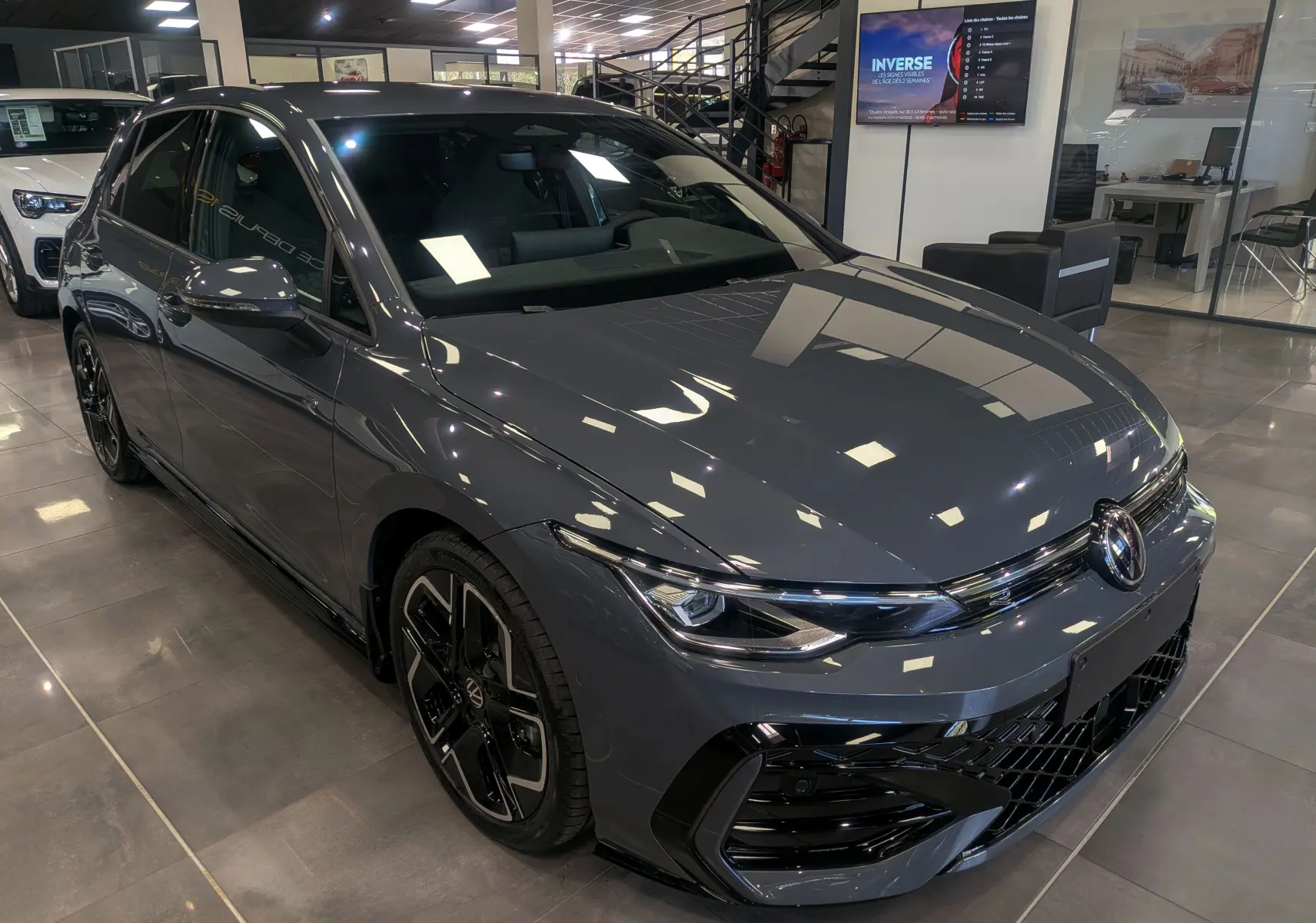 Vue 3/4 avant droite d'une Volkswagen Golf VIII 1.5 eTSI R-Line gris Urano avec jantes noires biton visibles en showroom.