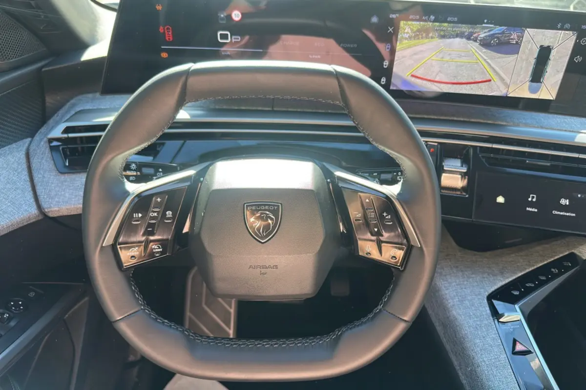 Vue intérieure centrée sur le volant cuir multifonctions du Peugeot 5008 gris, avec écran numérique affichant la caméra 360° en arrière-plan.