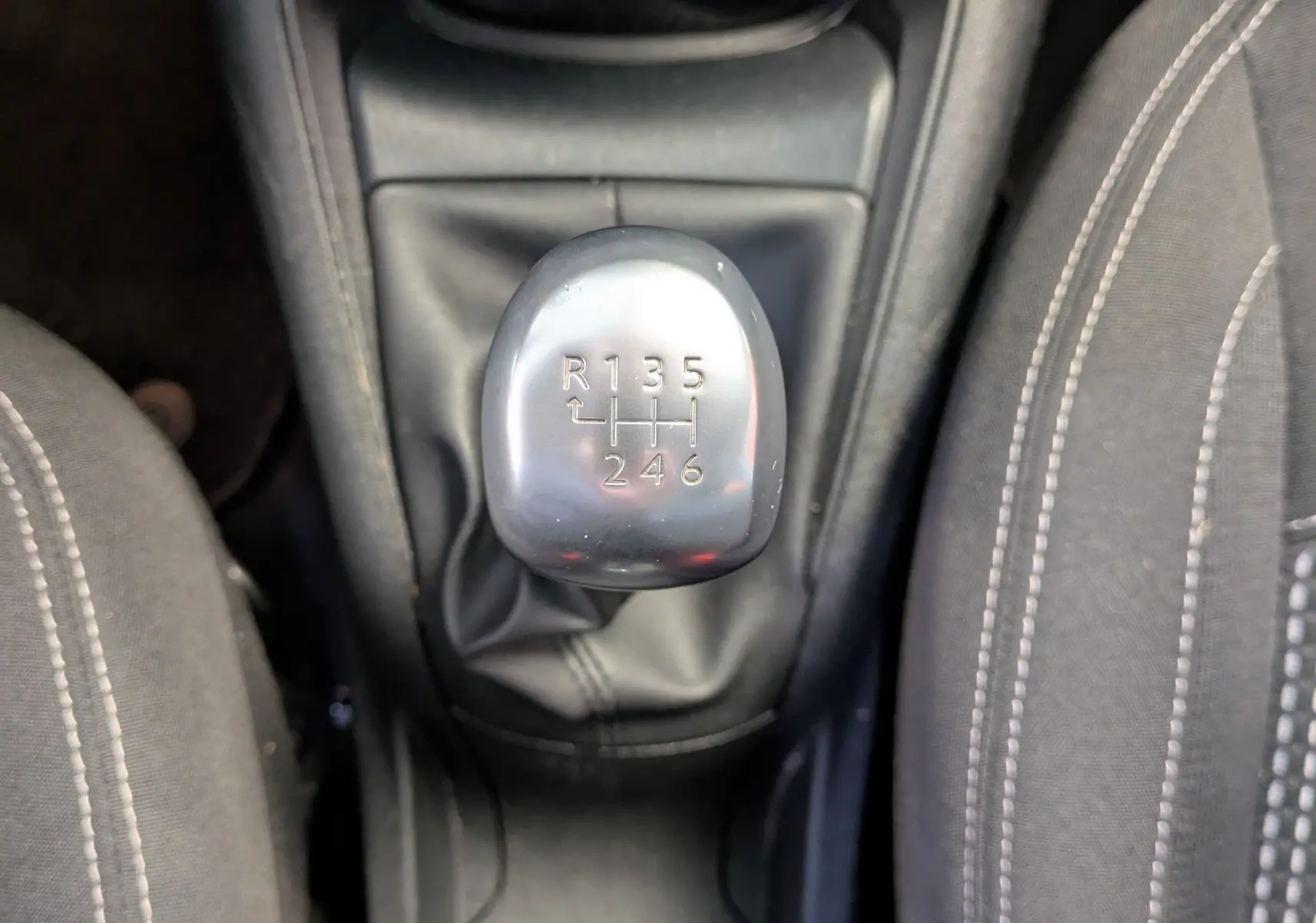 Vue plongeante sur le levier de vitesses manuel argenté entouré de sièges noirs avec surpiqûres blanches dans une Peugeot 208 noire.