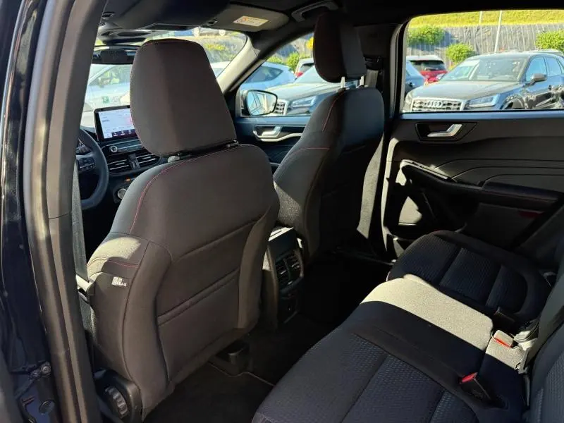 Vue intérieure arrière côté gauche du Ford Kuga 2.0 EcoBlue 2024, sièges noirs avec surpiqûres rouges et tableau de bord visible.