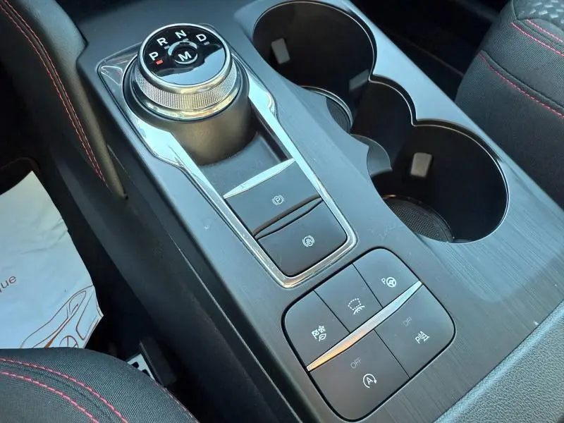 Gros plan sur la console centrale du Ford Kuga 2024 avec molette de sélection de vitesse et boutons de contrôle, intérieur noir avec surpiqûres rouges.