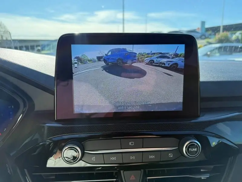 Vue écran caméra de recul montrant un Ford Kuga bleu Abysse en 3/4 arrière sur un parking ensoleillé.