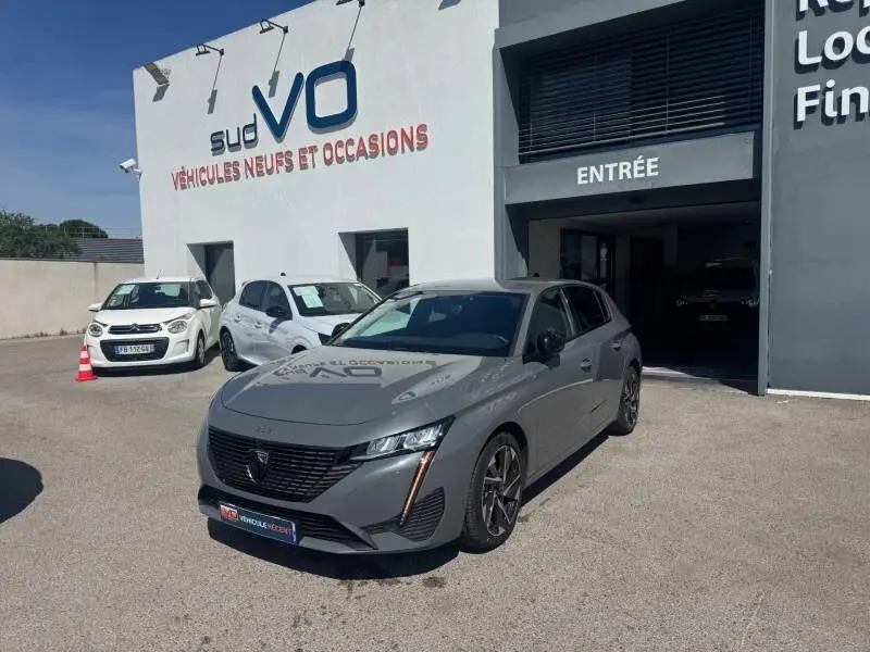 Peugeot 308 Hybrid gris vue 3/4 avant droit, jantes alliage 17 pouces, stationnée devant un garage.