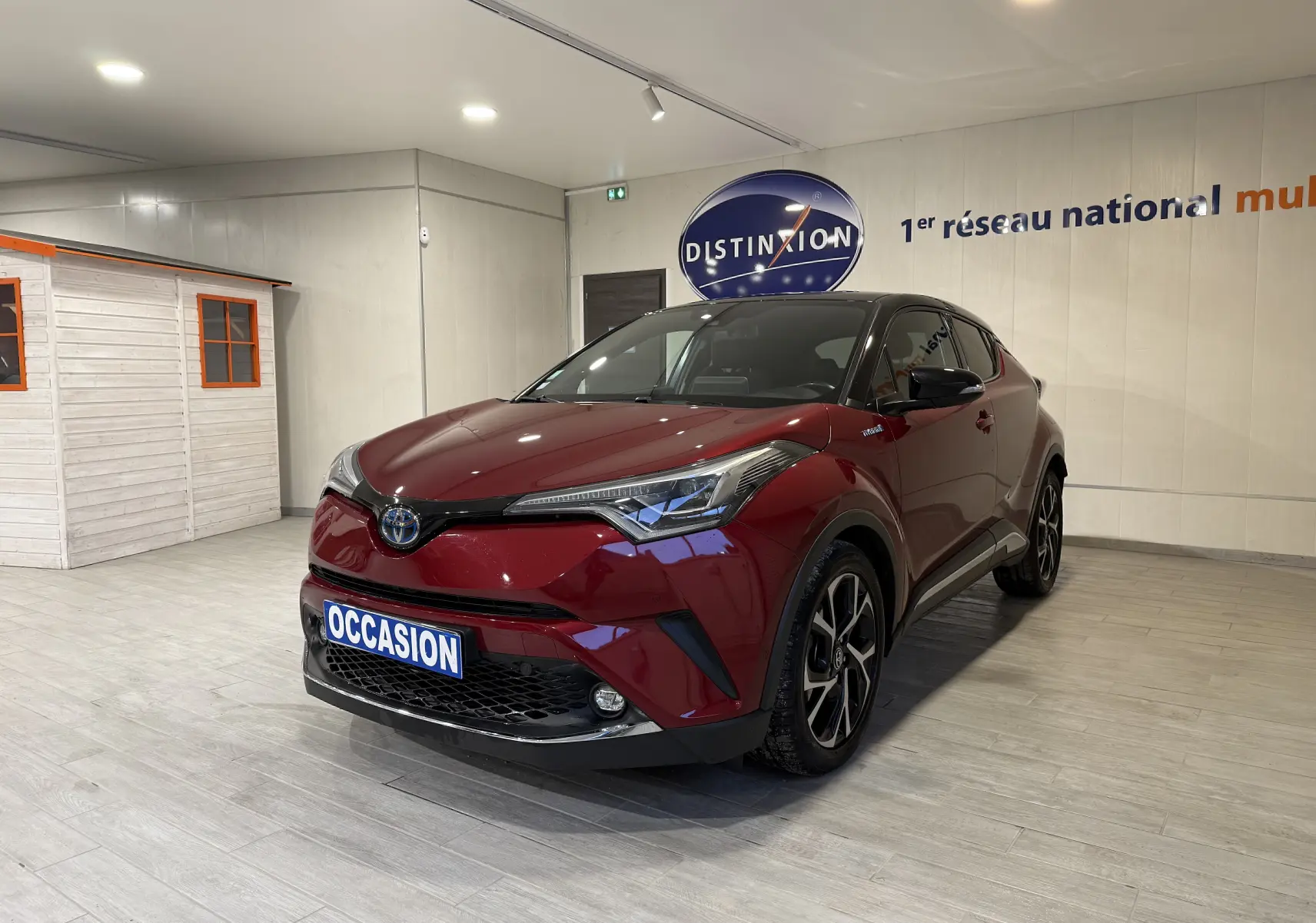 Toyota C-HR Hybride rouge en 3/4 avant droit, avec jantes noires et toit noir, présenté en intérieur showroom.