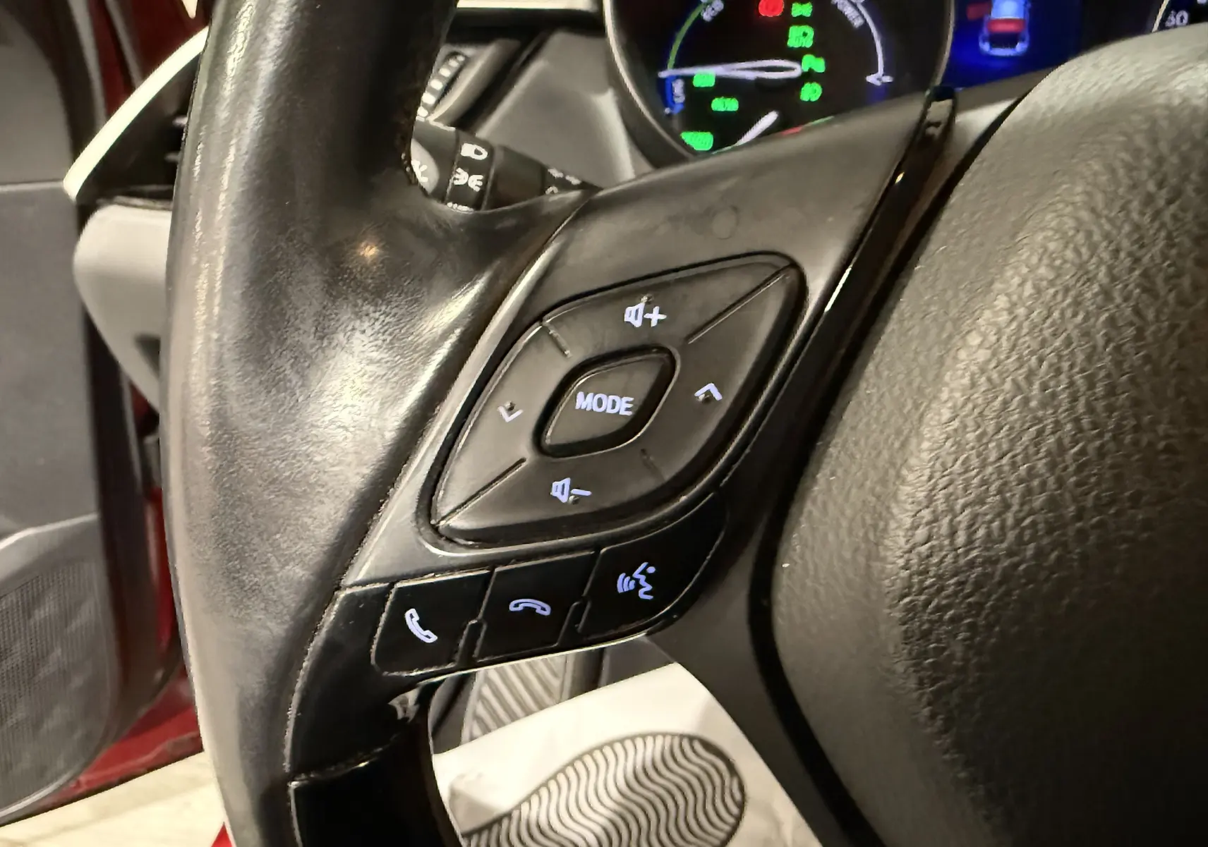 Gros plan sur les commandes du volant noir du Toyota C-HR Hybride rouge, avec tableau de bord numérique en arrière-plan.