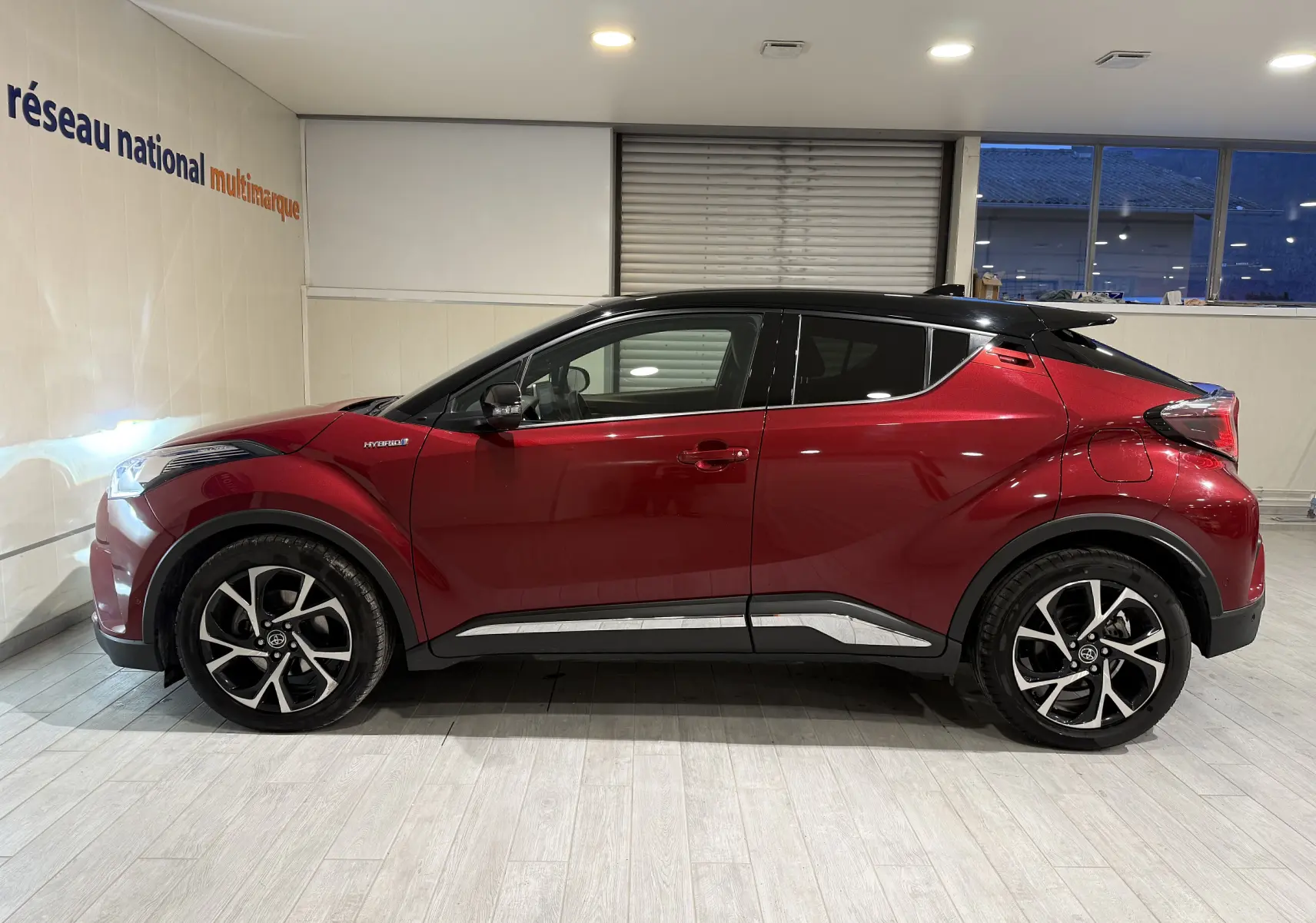 Vue de profil côté gauche d'un Toyota C-HR Hybride rouge avec toit noir et jantes alliage bicolores en intérieur showroom.
