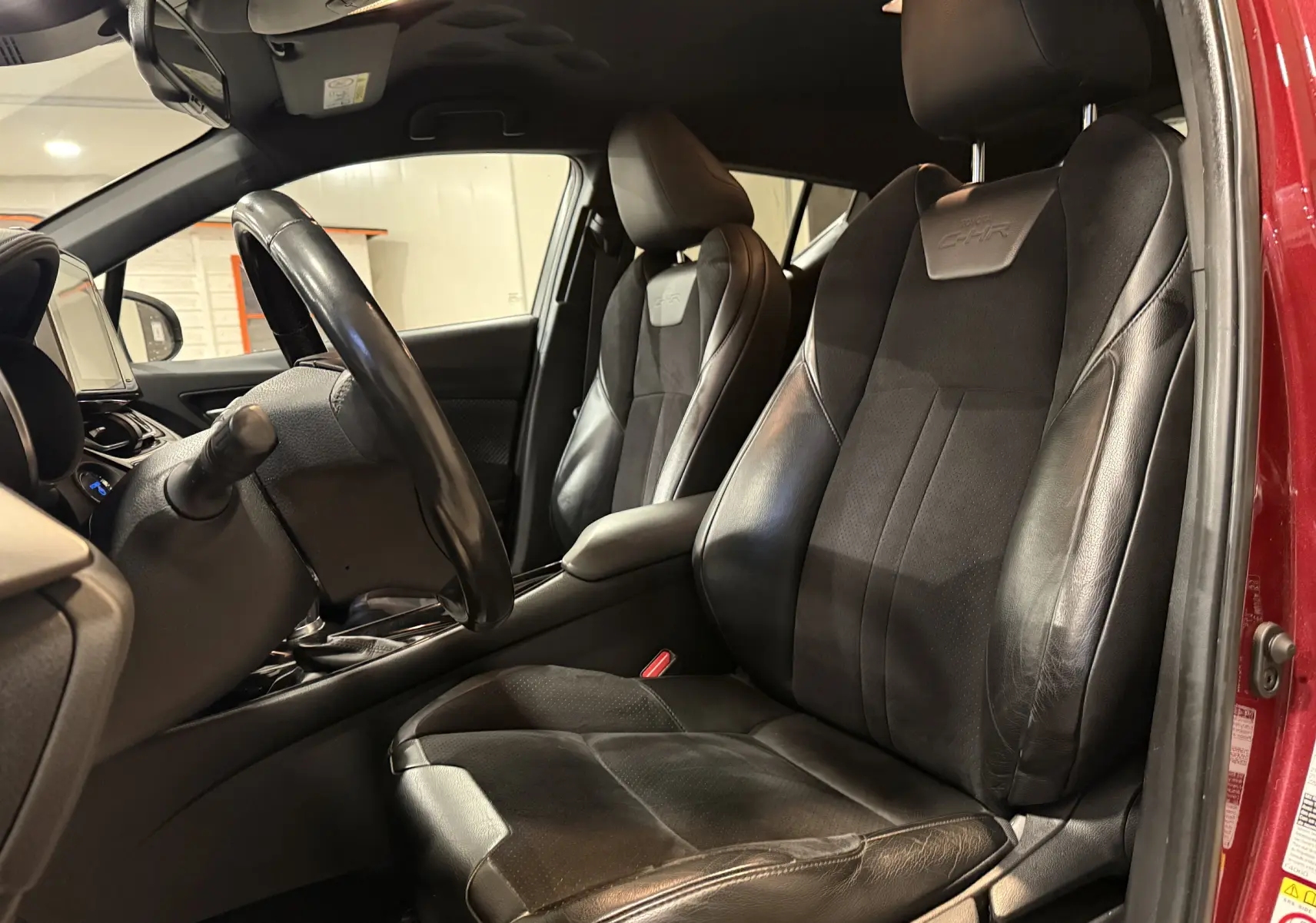 Vue intérieure côté conducteur du Toyota C-HR Hybride rouge, sièges en cuir noir avec logo C-HR embossé.