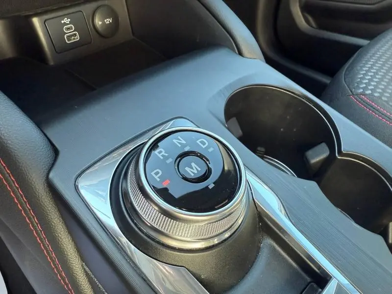 Gros plan sur la molette de sélection de vitesses noire et argentée dans l’habitacle du Ford Kuga bleu, avec porte-gobelets et prises USB visibles.
