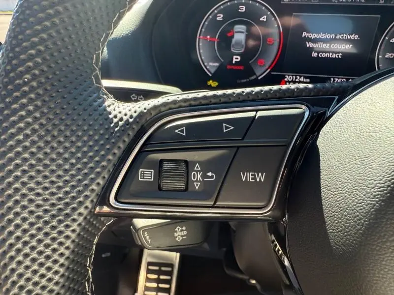 Gros plan sur les commandes au volant à gauche de l'Audi Q2 35 TDI 150ch S line 2025 avec affichage numérique en arrière-plan.