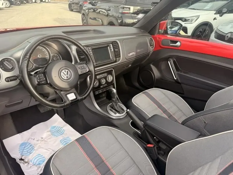 Vue intérieure côté conducteur de la Volkswagen Coccinelle Cabriolet rouge, avec sellerie grise et volant multifonction.
